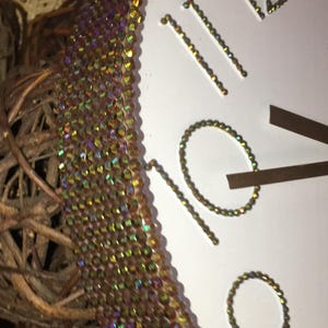 May include: A white clock face with gold-colored numerals and hands. The clock's edge is adorned with iridescent, multi-colored crystals. The clock is partially visible, with the numbers 9, 10, 11, and 12 in view.
