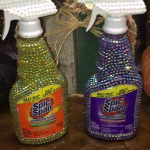 May include: Two bedazzled Spic Span spray bottles. One is covered in green rhinestones, the other in purple. Both have white spray nozzles and the text "KILLS 99.9% COVID-19" and "Spic Span Advanced Clean".