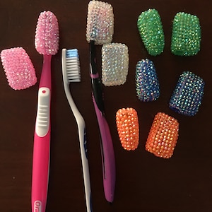 May include: A collection of toothbrushes and decorative toothbrush head covers. The toothbrushes are pink, purple, and white. The covers are embellished with sparkling rhinestones in pink, green, blue, orange, and iridescent colors.