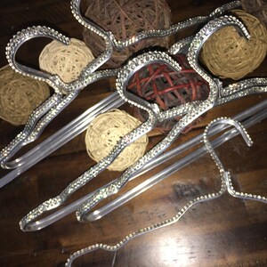 May include: A set of silver-toned metal clothing hangers featuring a decorative border of clear, sparkling rhinestones along the top edge and hook. The hangers have a wavy, contoured shape designed for garment support and display.