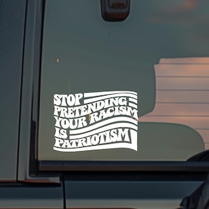 K&ouml;nnte beinhalten: Wei&szlig;er Vinyl-Aufkleber auf einer schwarzen Fahrzeugscheibe. Der Aufkleber lautet "STOP PRETENDING YOUR RACISM IS PATRIOTISM" in einer stilisierten Schriftart mit einem Flaggen-Design. Das Fahrzeug ist dunkel.