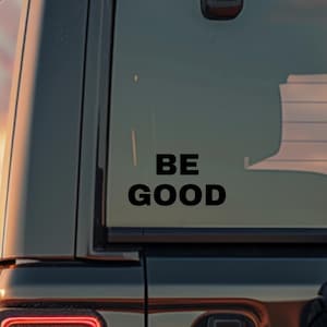 May include: Black vehicle with a rear window sticker that reads "BE GOOD" in a bold, sans-serif font. The vehicle's taillight is illuminated, and a spare tire is mounted on the back. The background shows a sunset.