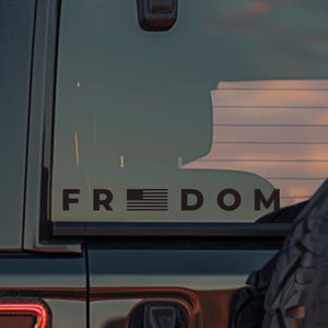 以下が含まれることがあります： 黒い「FREEDOM」デカール、黒いSUVの後部窓にアメリカ国旗のデザイン。デカールは太字のサンセリフ体です。背景には夕焼けが見えます。