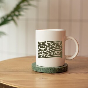 K&ouml;nnte beinhalten: Wei&szlig;e Keramik-Tasse mit dem Text "STOP PRETENDING YOUR RACISM IS PATRIOTISM" in gr&uuml;ner Schrift. Die Tasse steht auf einem gr&uuml;nen Marmor-Untersetzer auf einem Holztisch.