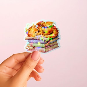 May include: A holographic sticker featuring a small orange dragon sleeping on a stack of four books. The dragon has closed eyes and small wings. The books are various colors including purple, green, and blue, with a small scroll resting on the bottom.