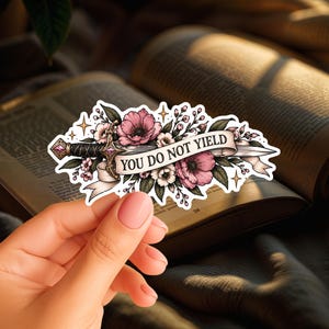 May include: A die-cut sticker featuring a silver sword intertwined with pink flowers and green leaves. A white banner draped across the blade displays the black text 'YOU DO NOT YIELD'. The design includes small gold star accents and a white border.