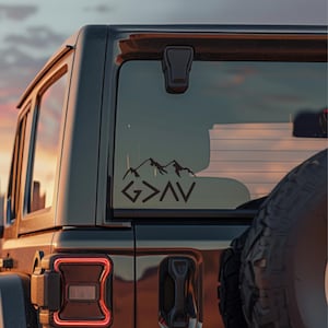 May include: Black vinyl decal with the text "G>AV" and a mountain range design, affixed to the rear window of a black SUV. The decal is visible against a sunset backdrop, showcasing a minimalist aesthetic.