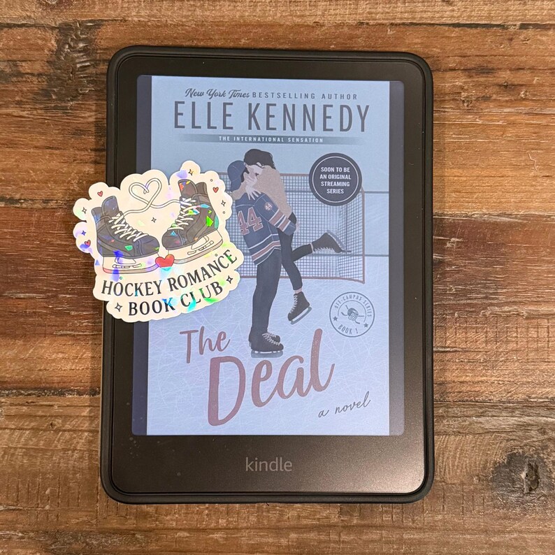 Op de afbeelding: Een Kindle e-reader met de cover van "The Deal" van Elle Kennedy, met een sticker met de tekst "Hockey Romance Book Club". De boekomslag toont een omhelzend stel. De e-reader ligt op een houten oppervlak.