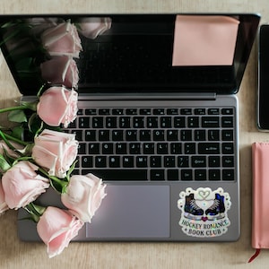 Op de afbeelding: Een laptop met een sticker met de tekst "Hockey Romance + Book Club", een roze notitieboek en een smartphone. Roze rozen zijn naast de laptop gerangschikt. Een roze plakbriefje zit op het scherm.