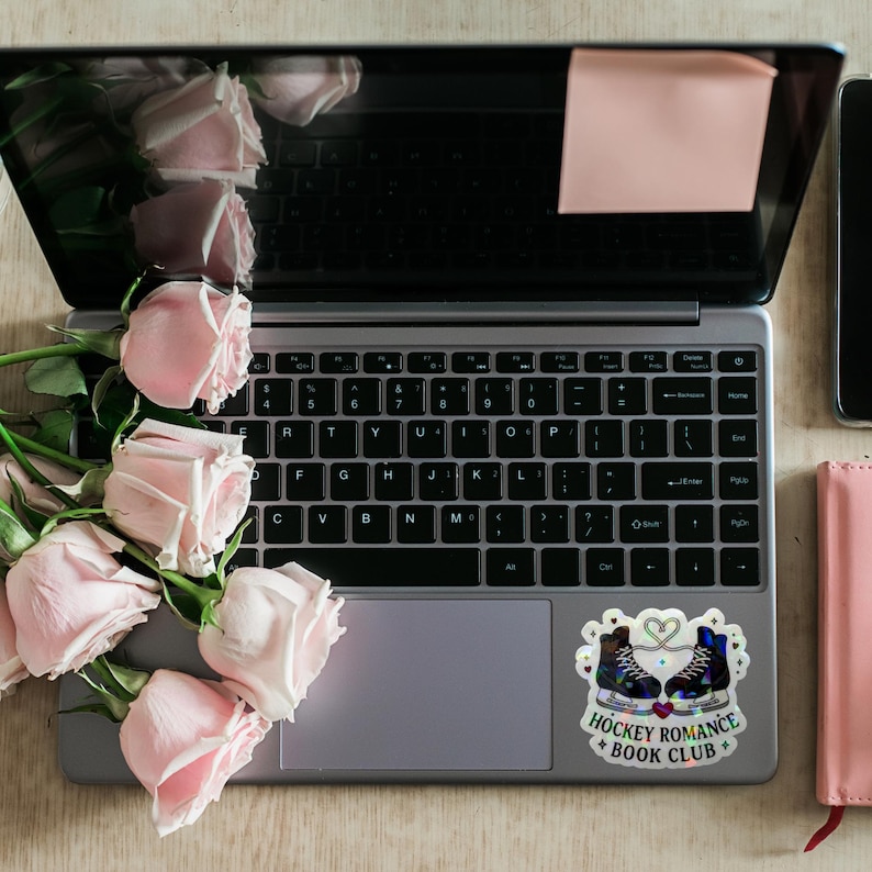 Op de afbeelding: Een laptop met een sticker met de tekst "Hockey Romance + Book Club", een roze notitieboek en een smartphone. Roze rozen zijn naast de laptop gerangschikt. Een roze plakbriefje zit op het scherm.