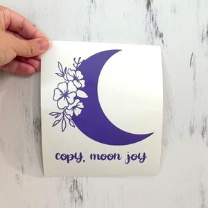 May include: A purple crescent moon sticker with floral accents and the words "copy, moon joy" in a script font. The sticker is held by a hand against a white wooden background.