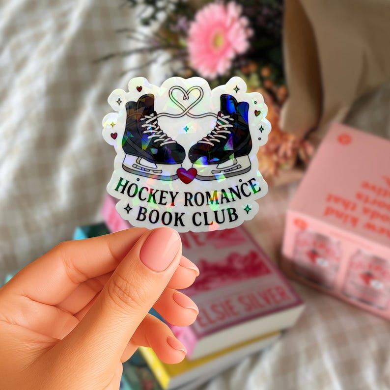 Op de afbeelding: Holografische sticker met twee schaatsen verbonden door een hart. De tekst "HOCKEY ROMANCE BOOK CLUB" staat eronder, met ster- en hartdetails. De sticker heeft een reflecterende, iriserende afwerking.