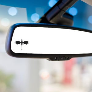 May include: A black vinyl decal of a dragon silhouette applied to the surface of a rectangular vehicle rearview mirror. The dragon is positioned on the left side of the mirror, featuring outstretched wings and a long tail with a fin at the end.