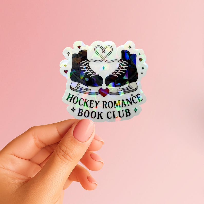 Op de afbeelding: Een holografische sticker met twee schaatsen tegenover elkaar, verbonden door een hart. De tekst "HOCKEY ROMANCE BOOK CLUB" staat eronder, met sterren. De sticker heeft een roze achtergrond.
