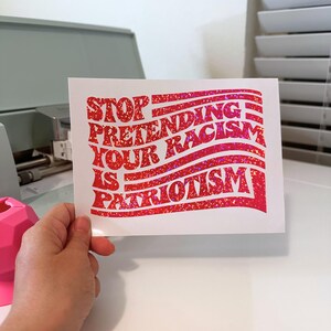 K&ouml;nnte beinhalten: Ein wei&szlig;er, rechteckiger Aufkleber mit rotem Glitzertext. Der Text lautet "STOP PRETENDING YOUR RACISM IS PATRIOTISM" in einem wellenf&ouml;rmigen, flaggenartigen Design.