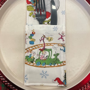 May include: A white fabric napkin with a green train and characters from the book "How the Grinch Stole Christmas!" printed on it. A fork and knife are tucked inside the napkin.