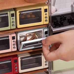Op de afbeelding: Een set van vijf miniatuurovens in verschillende kleuren, waaronder geel, groen, roze, rood en wit, zijn gestapeld op een houten plank. De ovens hebben glazen deuren en zijn verticaal in een rij gerangschikt. Een hand opent de deur van de lichtblauwe oven, waardoor een klein dienblad met twee stukjes voedsel zichtbaar wordt.