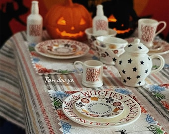 May include: A miniature Halloween-themed table setting with a carved pumpkin, plates, cups, and a teapot. The plates have Halloween-themed text and graphics. The teapot is white with black star patterns. The table is set with a striped tablecloth and spiderweb placemats.