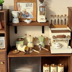 Könnte beinhalten: Ein Miniatur-Kaffee-Set mit einer weißen Espressomaschine, einem Mahlwerk, einem Kaffeebohnenspender und drei Glasröhrchen gefüllt mit Kaffeebohnen. Das Set enthält auch ein gerahmtes Poster mit dem Text "Das Leben kommt mit Kaffee" in Japanisch und Englisch, sowie drei weiße Keramik-Kaffekannen.