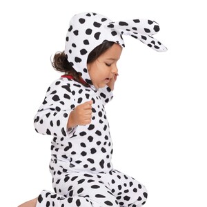 Könnte beinhalten: Ein Kind in einem Dalmatinerkostüm, bestehend aus einem weißen Outfit mit schwarzen Punkten und einer passenden Kapuze mit Schlappohren. Das Kind kniet auf weißem Hintergrund.