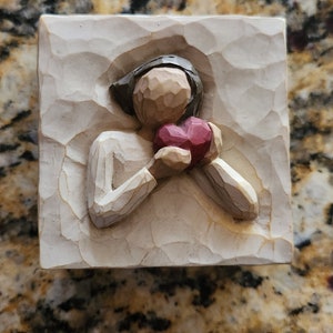 May include: A white wooden figurine of a person holding a red heart. The figurine is carved with a textured surface.