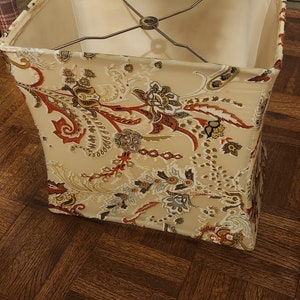 May include: A square lampshade with a beige background and a paisley pattern in shades of red, brown, and gold. The lampshade has a metal frame.