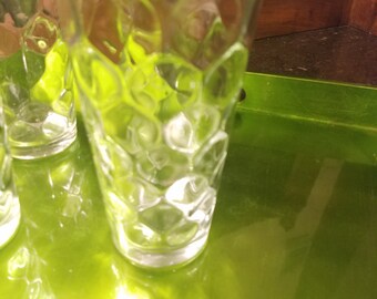 May include: Clear, textured drinking glasses with a honeycomb pattern. The glasses are on a bright green tray, reflecting light. The glasses are cylindrical with a slightly flared top.