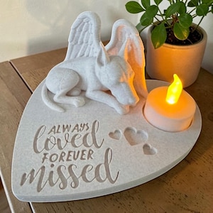 Könnte beinhalten: Eine weiße, herzförmige Gedenktafel mit einem skulptierten Hund mit Engelsflügeln. Der Text "ALWAYS loved FOREVER missed" ist zusammen mit drei kleinen Herzausschnitten und einer runden Aussparung für ein leuchtendes LED-Teelicht eingraviert.