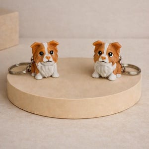 Op de afbeelding: Twee identieke sleutelhangers met een kleine, gebeeldhouwde hond met bruine en witte vacht, donkere ogen en een witte borst. Elk figuurtje is bevestigd aan een ronde, zilverkleurige metalen ring voor sleutels met een gedetailleerd ontwerp.