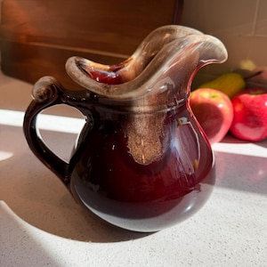 May include: A ceramic jug featuring a deep burgundy base that transitions into a lighter, creamy tan glaze around the wide, curved spout. The vessel has a rounded body and a sturdy, arched handle, showcasing a glossy, smooth finish throughout.
