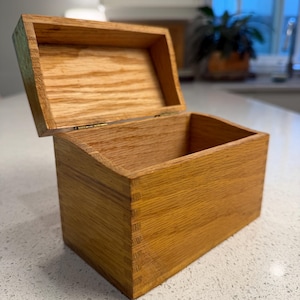 May include: A rectangular wooden box with a hinged lid, featuring a natural light brown wood grain finish and visible finger joint construction at the corners. The box is open, revealing a hollow interior and a smooth, light-toned wooden surface.