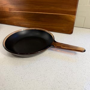 May include: A round frying pan featuring a matte black interior cooking surface and a translucent amber-coloured glass exterior. The pan includes a matching amber glass handle extending from the side, creating a distinct contrast between the dark basin and the warm, see-through glass rim and handle.