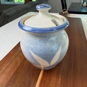 May include: A round ceramic jar with a matching lid featuring a small handle and a side notch. The jar has a speckled light blue and white glaze with a darker blue rim and a white leaf-like pattern around the base of the body.