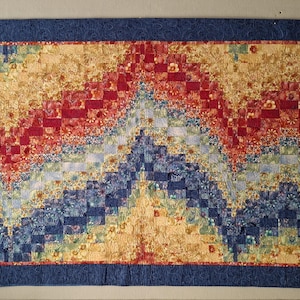 May include: A rectangular quilt with a blue border and a colorful patchwork design. The quilt features a central pattern of red, yellow, and blue blocks, creating a textured, layered effect. The design resembles a mountain range.