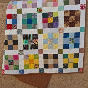 May include: A handmade patchwork quilt with a white background and squares of various colors and patterns. The quilt features squares of red, blue, green, yellow, and brown fabrics. The quilt is trimmed with a brown border.