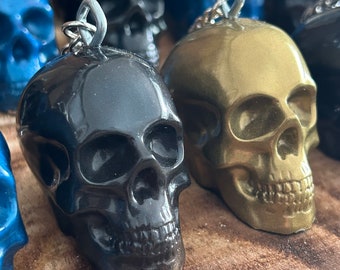 May include: Close-up of several skull-shaped keychains in various colors, including black, gold, and blue. Each keychain has a silver-colored loop for attaching to keys. The skulls are detailed and have a glossy finish, suggesting they are made of resin or a similar material.
