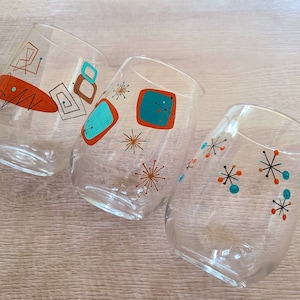 May include: Three clear glass drinking glasses with a mid-century modern design. The glasses are decorated with orange, turquoise, and black geometric shapes and starbursts.