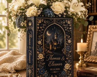 May include: A dark blue rectangular book-shaped box featuring gold foil illustrations of a castle, a crescent moon, and floral patterns. The text reads Thomas & Eleanor and 09.14.2026 on the front, with Thomas & Eleanor on the spine.