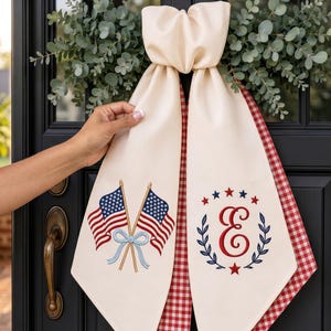 May include: A cream-colored fabric door hanger featuring a large bow at the top and two long tails. One tail displays two crossed American flags with a light blue ribbon, while the other features a red embroidered letter E surrounded by stars and leaves.
