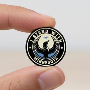 May include: A round black and white pin with the text "I STAND WITH MINNESOTA" encircling a central graphic of a bird with outstretched wings. The bird is black with blue accents, and a star is above it.