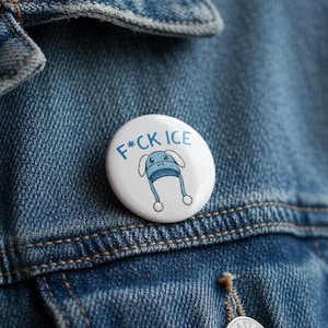 Könnte beinhalten: Ein weißer Ansteckbutton mit dem blauen Text "F*CK ICE" und einem Cartoon-Hut-Design. Der Button ist an einer blauen Jeansjacke befestigt.