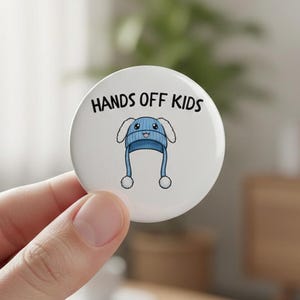 May include: A white button with the text "HANDS OFF KIDS" in black. The button features a cartoon illustration of a blue knit hat with white ear flaps and two white pom-poms. The button is round.