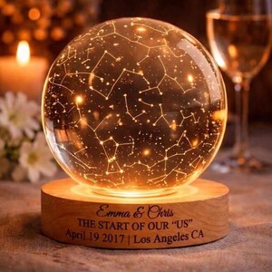 Op de afbeelding: Een verlichte glazen bol met sterrenbeelden, geplaatst op een houten voet. De voet is gegraveerd met de namen "Emma & Chris", de zin "THE START OF OUR US", de datum 19 april 2017 en "Los Angeles CA".