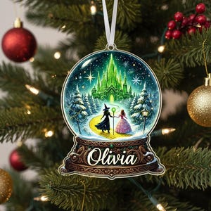 May include: A personalized Christmas ornament featuring a snow globe design with a fantasy scene. The ornament shows a green castle, two figures, and the name "Olivia" in a decorative font. It hangs from a white ribbon on a Christmas tree.