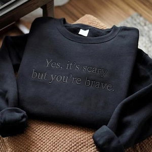 Op de afbeelding: Een zwart sweatshirt met de tekst "Yes, it's scary but you're brave" geborduurd op de voorkant. Het sweatshirt ligt op een bruin geweven oppervlak. De mouwen zijn opgerold bij de polsen.
