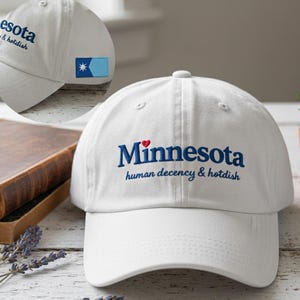 May include: White baseball cap with the word "Minnesota" embroidered in navy blue with a red heart above it. The phrase "human decency & hotdish" is embroidered below. A small flag patch is on the side.
