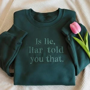 Può includere: Una felpa verde scuro con la scritta ricamata "Is lie, liar told you that". Un tulipano rosa con stelo e foglie verdi poggia sulla felpa. La felpa è piegata su una coperta color crema.