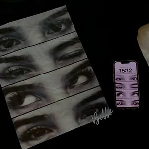 以下が含まれることがあります： 黒いTシャツ。複数の目が並んだグラフィックが特徴です。デザインには右下にサインが含まれています。スマートフォンには同様の目のグラフィックが表示されています。電話の時刻は15:12です。