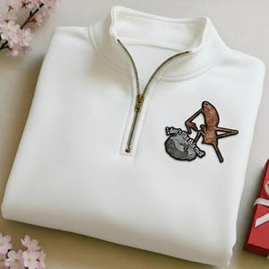 Pode incluir: Uma camisola branca com fecho de correr a um quarto e fecho de correr dourado. A camisola apresenta um patch com o texto "I don't like this rock" e um gráfico de uma criatura e uma rocha. Uma caixa de presente vermelha com uma fita branca está no fundo.