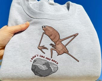 May include: A light gray sweatshirt featuring a cartoonish creature with long limbs and the text "I DON'T LIKE THIS ROCK." The graphic includes a gray rock. The sweatshirt is made of a soft material.